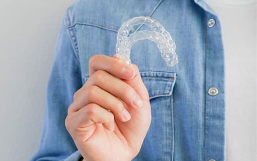 Person wearing a jean jacket while holding up clear aligners with one hand.